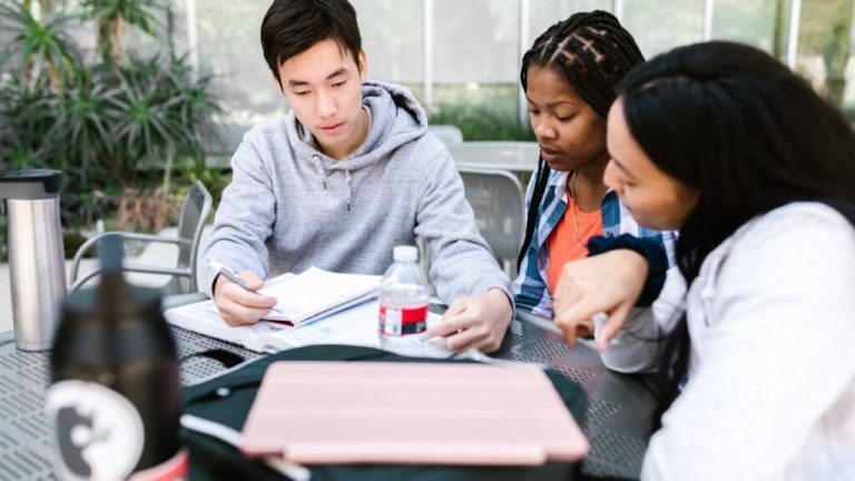 Three students, two women and one man, studying together at an outdoor table with books and a laptop.