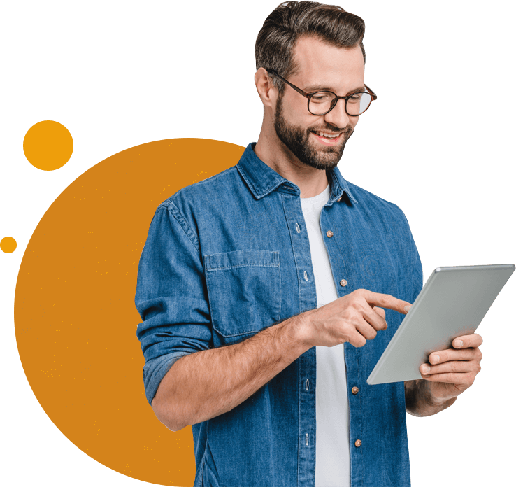 A smiling man in a denim shirt using a tablet, standing against a background with orange circles, possibly browsing overseas education consultants.