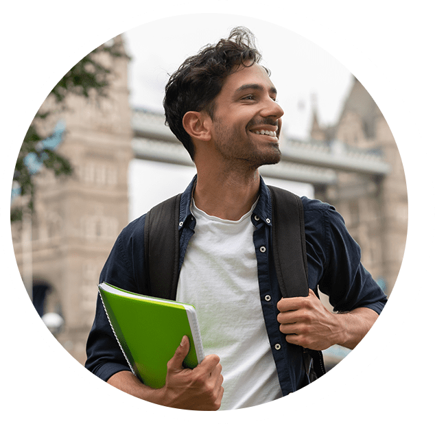 A smiling young man with a backpack and a green notebook looks up, standing near a bridge in an urban setting.