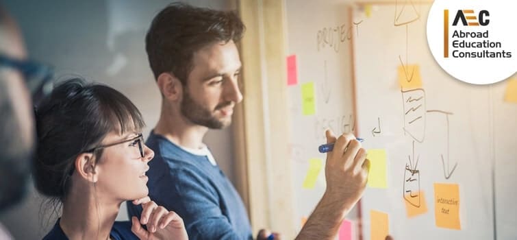 Two professionals, a man and a woman, discussing and writing on a whiteboard with sticky notes in an office setting. the logo of abroad education consultants is visible.