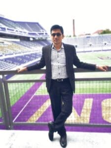 Man in a black blazer and checkered shirt standing with arms spread wide in front of stadium seating.