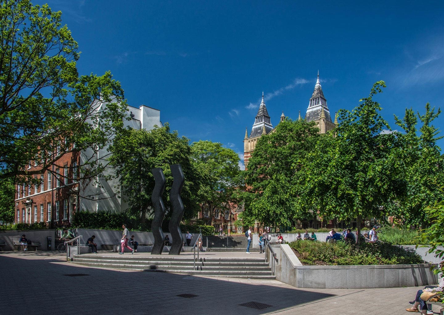 An urban park near the University of Leeds with people relaxing, featuring modern sculptures, steps, surrounded by lush trees, and distant gothic-style towers.