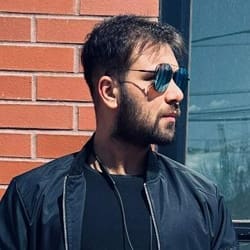 A man with a beard wearing sunglasses and a black jacket stands outside near a brick wall, looking to the side.