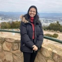 A woman in a black coat and red scarf smiling at the camera with a scenic city view in the background.