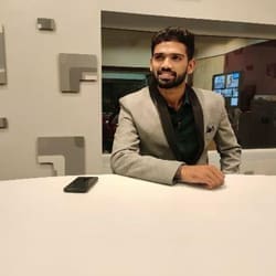 A young man in a gray blazer sitting at a table, smiling at the camera with a smartphone beside him.