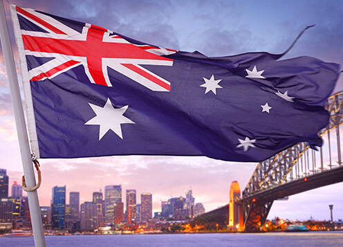 Australian flag waving in the foreground with the sydney skyline and harbour bridge in the background at dusk.