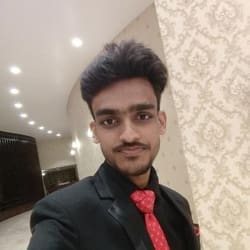 Young man in a black suit and red tie smiling for a selfie in a room with decorative wallpaper and ceiling lights.