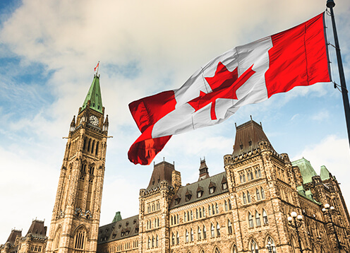 Canadian flag waving in front of the gothic-style parliament buildings under a partly cloudy sky.