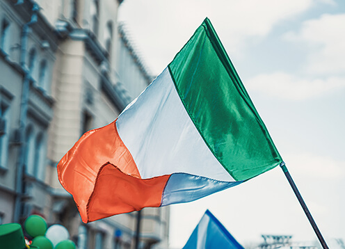 Irish flag waving in the wind against a backdrop of urban buildings, under a cloudy sky.