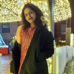 Woman standing outdoors at night, wearing a red top and black coat, with festive lights in the background.