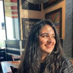 Young woman with long, dark curly hair smiling and looking away, seated in a restaurant with abstract art in the background.