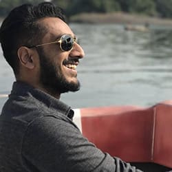 A man wearing sunglasses smiles broadly on a boat with water and trees in the background.