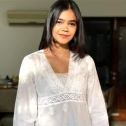 A young woman with shoulder-length black hair, smiling, wearing a white lace-detailed top, standing indoors with soft lighting.