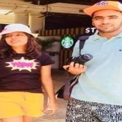 Two people, a woman in a white hat and a man holding a camera, smiling in front of a storefront.