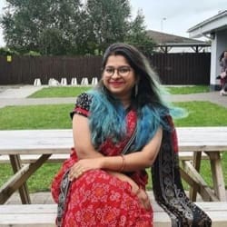 A woman with blue-tinted hair and glasses, smiling, sitting on a wooden bench in a park, wearing a red dress and a black shawl.