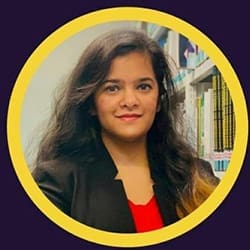 A professional woman with long dark hair, wearing a black blazer and red top, smiles gently in front of a bookshelf.