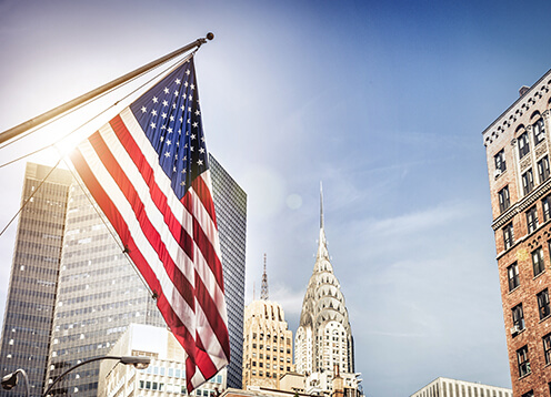 American flag waving in front of skyscrapers with the chrysler building visible in the background under a clear blue sky.