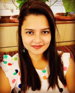 Young woman with shoulder-length brown hair smiling at the camera, wearing a white top with colorful polka dots, seated indoors.