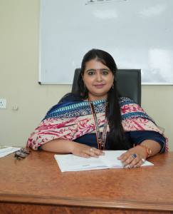 A woman seated at a desk in an office, smiling at the camera, wearing a patterned shawl over her outfit, with papers and a pen in front of her.