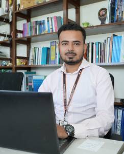 Man with a beard sitting at a desk with a laptop in an office, surrounded by bookshelves filled with books.