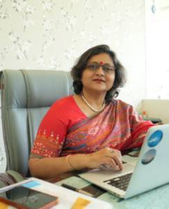 A woman in a red sari working on a laptop at a desk, with a smartphone and portable fan nearby.