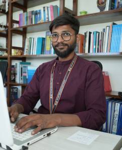 Man in a burgundy shirt and eyeglasses working at a computer in a library with books in the background.