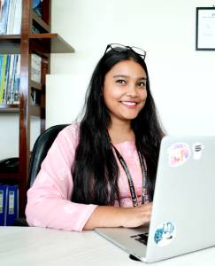 A young woman in a pink shirt and glasses smiles while working on a laptop in an office setting.