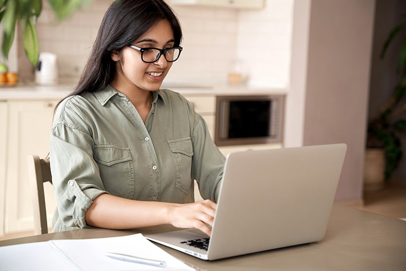 A woman wearing glasses and a green shirt smiles while using a laptop to study an IELTS Online Course at a kitchen table.